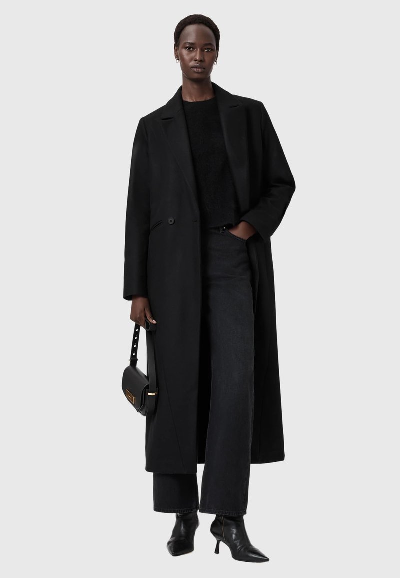 Cappotto lungo nero in lana con un solo bottone, abbinato a un maglione nero, jeans a gamba larga e stivaletti alla caviglia a punta; tiene una borsa nera.