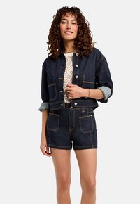 Ensemble en jean foncé comprenant une veste et un short, avec des coutures contrastées, de grandes poches et des poignets retroussés, surmonté d'un haut texturé blanc.