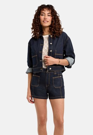 Set di giacca di jeans e pantaloni corti di colore blu scuro, con cuciture a contrasto, tasche oversize e maniche arrotolate, accompagnato da una maglietta bianca a trama sottostante.