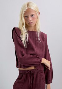 Top corto texturizado burdeos con mangas largas, que presenta un escote cuadrado y aberturas laterales. Combinado con pantalones de cintura elástica.