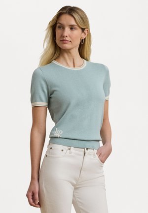 Lyseblå kortermet genser med ribbestrikket hals og kant, med hvit kanting og en brodert logo på den nedre siden. Bært med hvite jeans.
