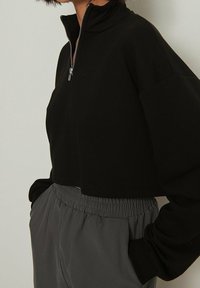 Schwarzes, kurzes Sweatshirt mit hohem Kragen und halbem Reißverschluss, kombiniert mit lockeren, grauen Hosen mit elastischem Bund. Weicher Stoff.