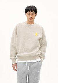 Jonge man in een beige sweatshirt met een gele sleutelgatlogo op de borst en grijze sweatpants met een witte koord en logo op het linkerbeen.