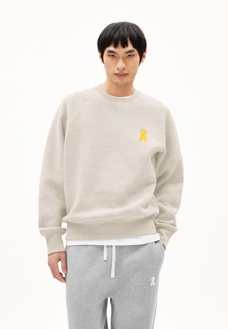 Jonge man in een beige sweatshirt met een gele sleutelgatlogo op de borst en grijze sweatpants met een witte koord en logo op het linkerbeen.