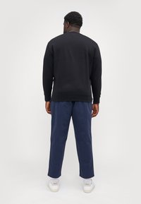 Black sweatshirt with long sleeves and crew neck, paired with navy blue pants, shown from the back. White sneakers visible at the bottom.