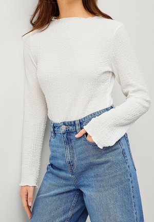 Kobieta w białej, teksturowanej bluzce z długim rękawem zakończonym lekko rozkloszowanymi mankietami oraz w niebieskich, wysokim stanem dżinsach, z ręką w kieszeni.