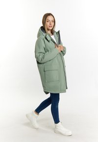 Grøn vandtæt parka med hætte, frontlynlås, to store lommer og kontrastforing, parret med blå jeans og hvide sneakers.