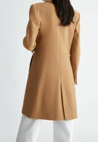 Kamelwollmantel mit einer maßgeschneiderten Passform, langen Ärmeln, Rückenschlitz und minimalen Details, kombiniert mit weißen Hosen.