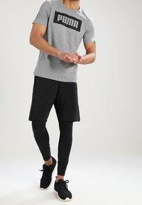 Harmaa lyhythihainen t-paita, jossa on suuri musta "PUMA" -logo, yhdistetty mustiin shortseihin ja tiukkapohjaisiin mustiin legginsseihin, käytetty mustien urheilukenkiin.