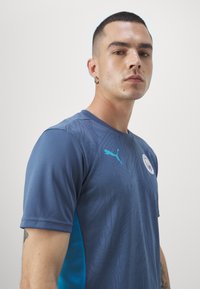 Gai zils sporta krekls ar teksturētu rakstu, īsām piedurknēm, Puma logo turkuza krāsā un apaļas kakla dizains.