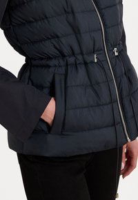 Marineblauwe pufferjack met gewatteerde textuur, een rits aan de voorkant en een koord in de taille. Beschikt over zijzakken en aangesloten zwarte mouwen.