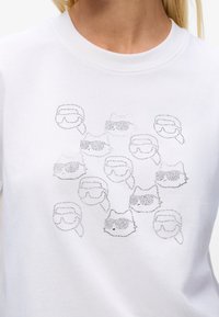 Weißes Shirt mit kleinen Strasssteinen in Form von Katzen und menschlichen Gesichtern, die Sonnenbrillen tragen, in einem verstreuten Muster auf der Brust.