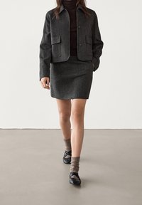 Dark gray tweed jacket with two front pockets, paired with a matching skirt. Model wears black shoes and ribbed brown socks.