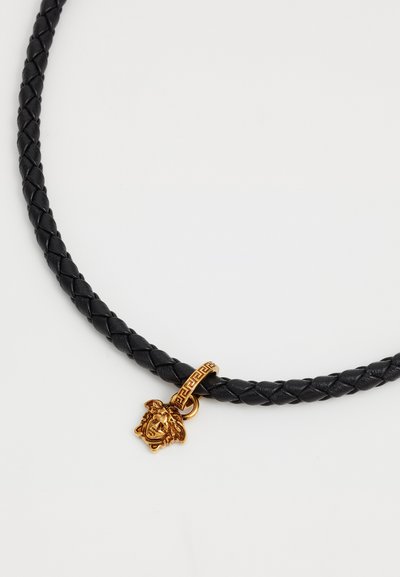 Črna pletena usnjena choker z zlatim obeskom v obliki leva in dekorativnimi zlatimi okrasnimi elementi. Oblikovanje vključuje teksturiran, tkani vzorec.