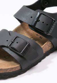 Musta nahkainen sandaali, jossa on kaksi säädettävää hihnaa, metalliset soljet ja korkkipohjallinen. Kuvioitu päällinen ja kumipohja pitävyyttä varten.