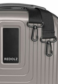 Zilverkleurige harde koffer met textuur, voorzien van een zwarte afneembare schouderriem met plastic draaiklemmen en een merklabel "REDOLZ".