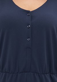 Navy blue jumpsuit with a V-neck, five button closure, textured fabric, and a gathered elastic waistband. Features side pockets.