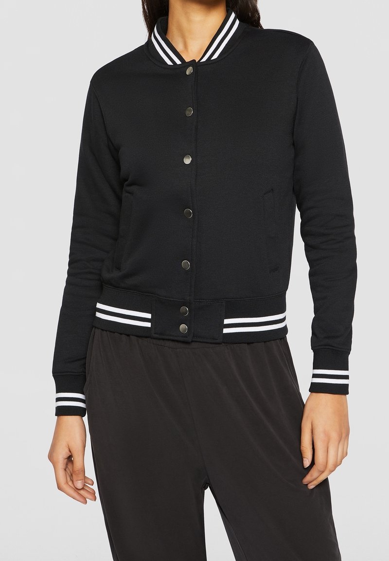 Femme portant une veste varsity noire avec un col, des poignets et une bande inférieure côtelés à rayures blanches et des boutons-pression noirs sur le devant.