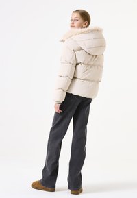 Leichte beige Daunenjacke mit Kapuze und Kunstfellbesatz, ausgestattet mit einem gesteppten Design und elastischen Bündchen. Kombiniert mit locker sitzenden dunklen Jeans.