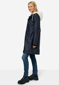 Námořnický voděodolný kabát s bílou kapucí a oranžovým podšíváním, vynikající přiléhavým střihem, se zapínacími kapsami a střední délkou.