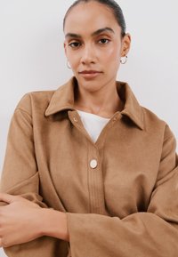 Ruskoinen, pehmeä kankainen takki, jossa on kaulus ja nappisuljenta. Ominaisuuksiin kuuluu leveät hihat ja sileä pinta, yhdistettynä valkoiseen paita alla.