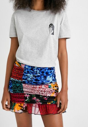Femme portant un t-shirt gris clair avec un petit graphique abstrait de visage et une mini-jupe colorée à motifs froncée avec un ourlet à volants.
