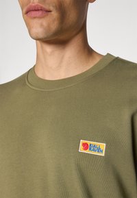 Olajzöld pamut póló kerek nyakkal. Négyzet alakú folttal rendelkezik piros és kék "Fjällräven" logóval, sárga háttérrel.