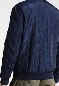 Marinblå vadderad bomberjacka tillverkad av glansigt nylon med ribbad midja och ärmslut. Har diamantformad sömnad och en slät yta.