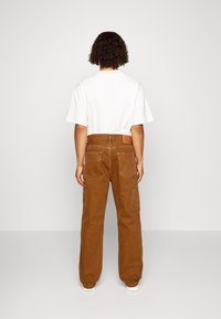 Pantalon cargo en toile marron avec plusieurs poches et un écusson logo à l'arrière. Associé à un t-shirt blanc à manches courtes.