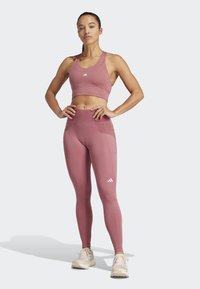 Mujer en un sujetador deportivo y leggings color malva con logos de Adidas, de pie con confianza con las manos en las caderas y usando zapatillas blancas.