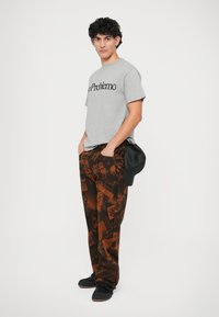 Graues Rundhals-T-Shirt mit dem Text "No Problemo", kombiniert mit braunen gemusterten Cargo-Hosen, die Text und Grafiken aufweisen. Schwarze Schuhe und eine Mütze.