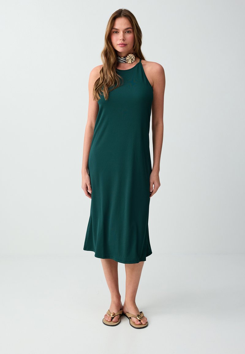 Femme portant une robe midi vert foncé sans manches avec un col halter et un collier ras du cou rayé avec une grande épingle décorative, debout devant un fond uni.