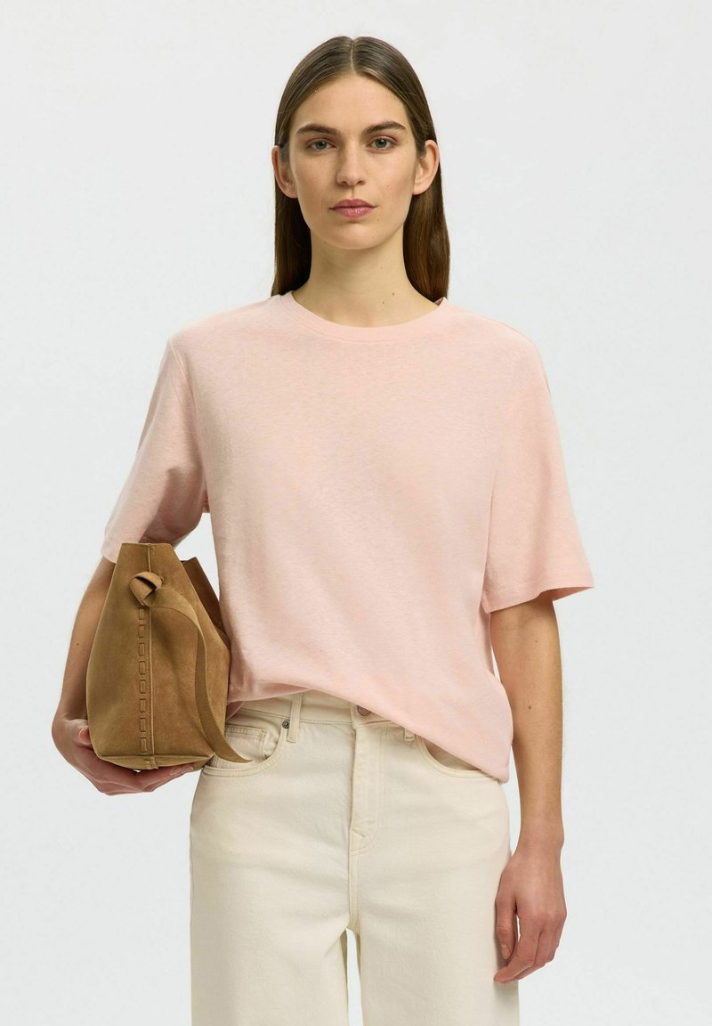 Femme aux cheveux bruns et raides portant un t-shirt rose pâle à manches courtes et un pantalon crème, tenant un sac à main en daim beige à ses côtés.