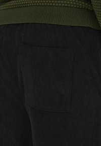 Schwarze Cordhosen mit einer Gesäßtasche und geripptem Material. Der Bund ist grün, akzentuiert durch ein gemustertes Strickdetail.