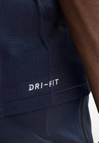 Haut de sport Dri-FIT bleu marine avec une texture en maille respirante ; présente un logo blanc proéminent près de l'ourlet, mettant en valeur son design sportif.