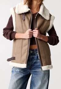 Gilet in pile marrone con colletto in pelliccia bianca, chiusura con zip e dettagli in pelle marrone. Presenta una vestibilità comoda e tasche laterali. Indossato sopra jeans di denim.