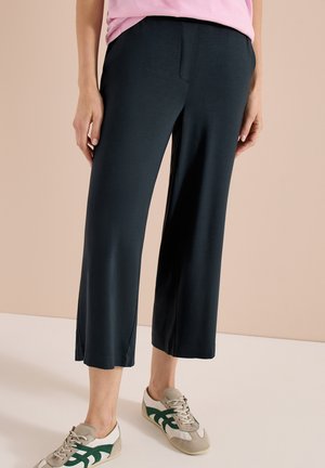 Persoon draagt zwarte wijde cropped broek, lichtroze top en beige sneakers met groen ontwerp, staand op een effen vloer.