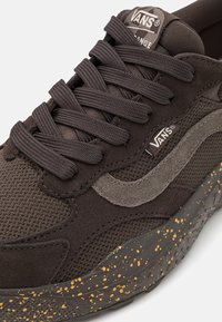 Sapatilhas Vans castanhas com um upper texturizado em camurça e rede, atacadores redondos, uma etiqueta de logótipo e uma sola de borracha salpicada em cinza escuro e amarelo.