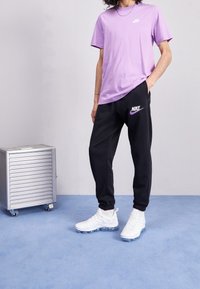 Lila bomullströja med en liten vit logo, kombinerad med svarta sweatpants som har en lila logo. Vita sneakers med klara sulor.