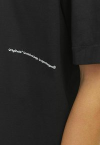 Sort kortærmet T-shirt lavet af bomuld, med et hvidt trykt logo på siden, der står "Originals™ Creative Dept. Copenhagen."