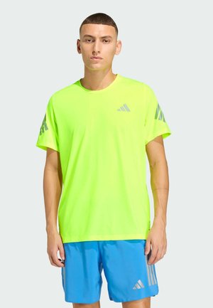 Camiseta deportiva de color amarillo neón con mangas cortas y cuello redondo, con detalles del logo de Adidas en gris en los hombros. Combinada con pantalones cortos azules.