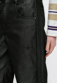 Crne traperice s blagim sjajem, s detaljima sa strane, prednjim džepovima i suptilnim crnim logom blizu bokova.