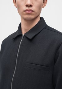 Jeune homme portant une veste noire zippée avec un col replié et une petite poche poitrine sur un fond clair uni.