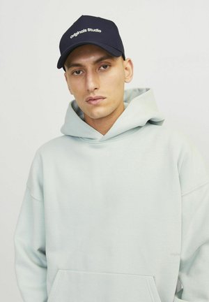 Hoodie vert menthe clair avec une poche kangourou, associé à une casquette de baseball bleu marine portant le texte "Originals Studio" en blanc. Texture lisse.