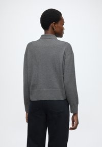 Pulover gri tricotat cu guler, design cropped, mâneci lungi și tiv cu dungi, asortat cu pantaloni negri, văzut din spate.