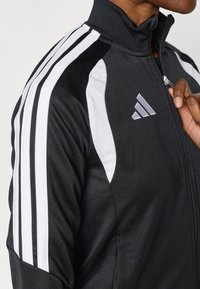 Μαύρο αθλητικό μπουφάν με λευκές ρίγες στους ώμους και ψηλό γιακά. Διαθέτει κλείσιμο με φερμουάρ και κεντη τό λογότυπο στο στήθος.