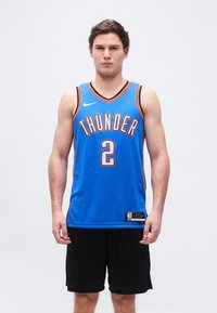 Jeune homme debout face à nous, portant un maillot de basket Thunder bleu avec le numéro 2 et un short noir.