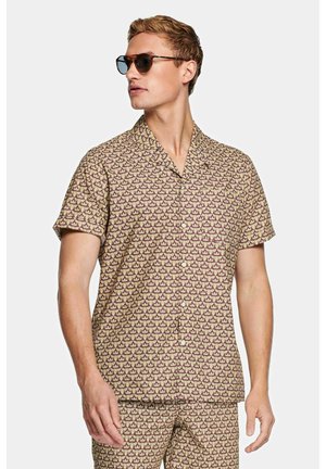 Man draagt een lichtbruin overhemd met korte mouwen en bijpassende korte broek met een patroon van kleine paarse en rode zeilboten, donkere zonnebril, kijkt naar links.