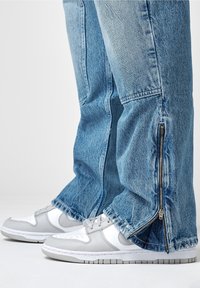 Světle modré džíny z denimu s zipem na lemech, spárované s bílými a šedými teniskami, které mají perforace a strukturovanou podrážku.