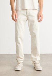 Man wearing off-white straight-leg jeans, beige suede sneakers, and a white shirt standing on a light gray floor against a white wall.